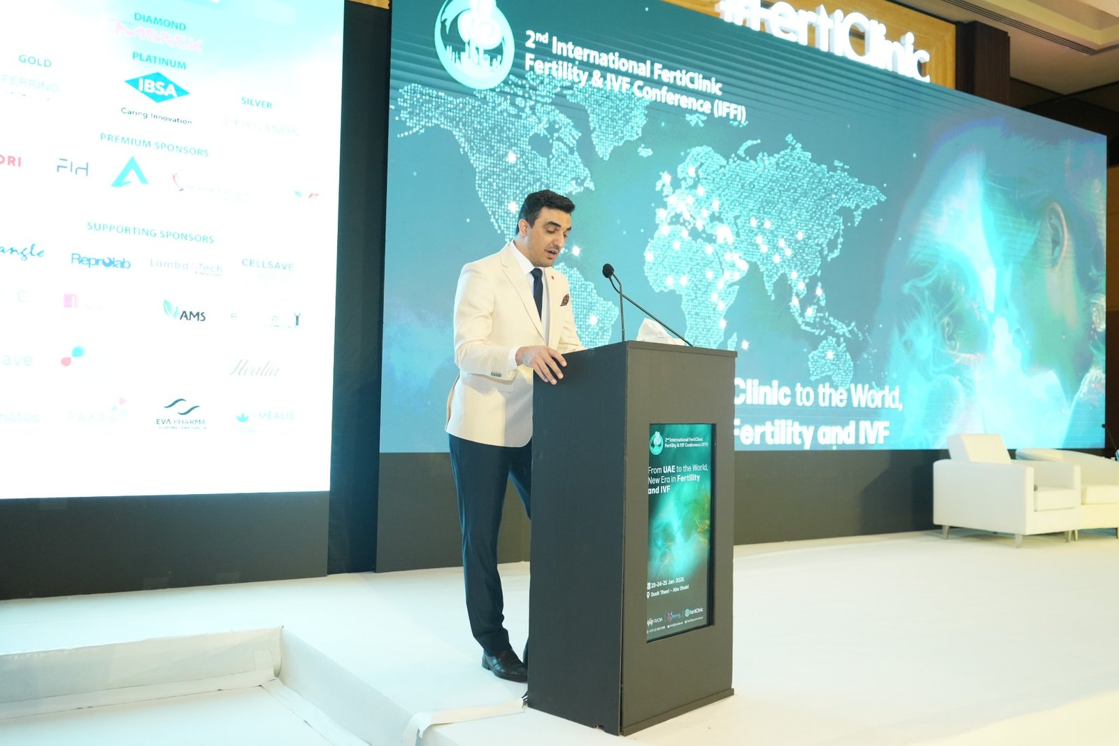 Dr. Alireza Amin in a cream jacket at the lectern, with conference and sponsor displays behind him.