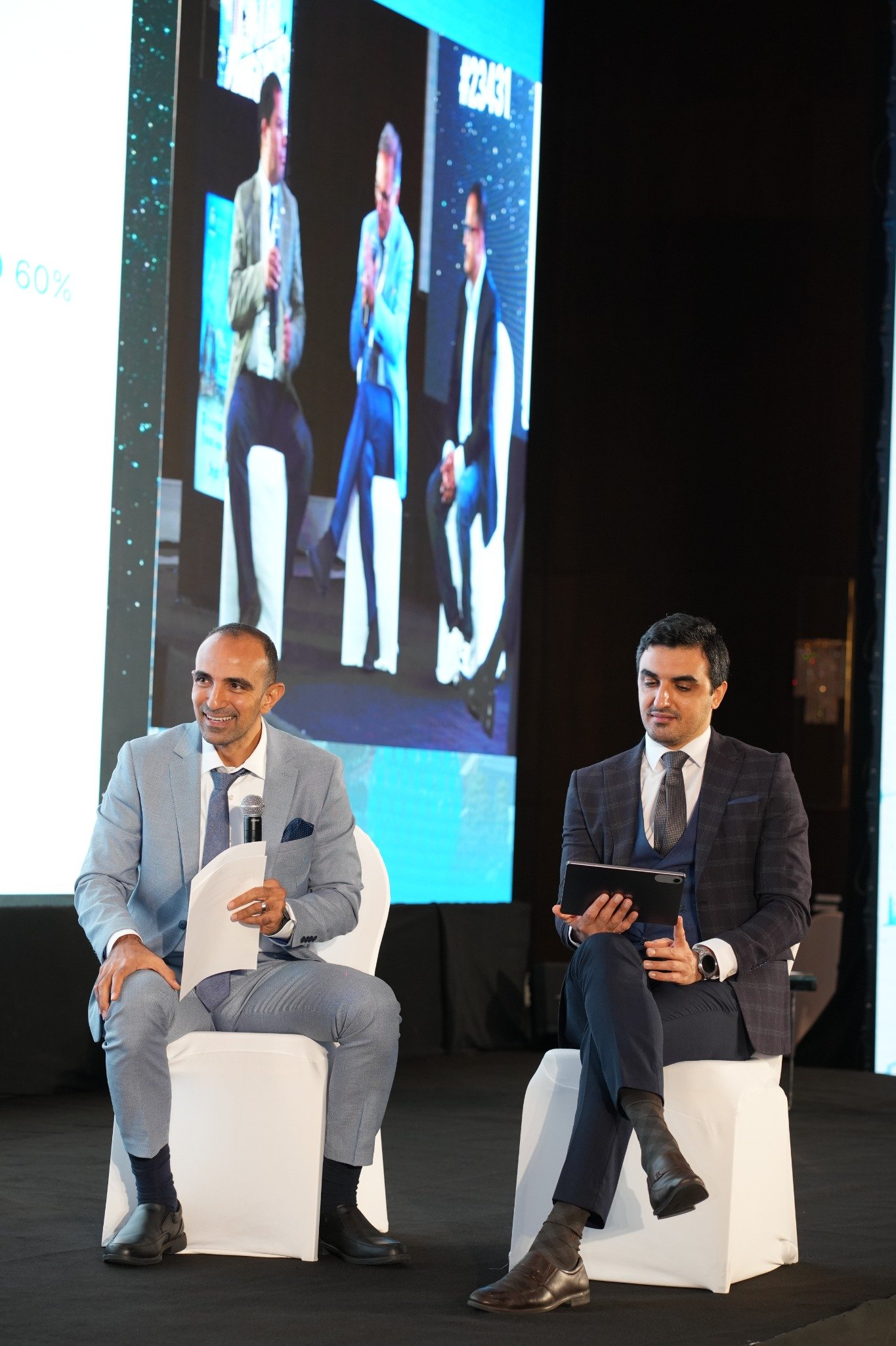 Two people seated on a conference stage with a microphone, papers and a tablet.